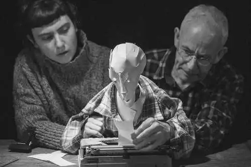 Schwarz weißes Bild: Ein junger und ein alter Mensch halten mit jeweils einer Hand eine Figur mit weißem Papierkopf. Die Figur schreibt auf einer Schreibmaschine.
