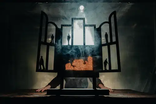 Schemenhafte Figuren vor einem transparenten Vorhang, der in unterschiedlichen Farben das Licht bricht. Im oberen Bereich ein Pferd neben einem Lichtkegel. Es wirkt wie eine Menschenansammlung, die vor einer lichtdurchfluteten Welle steht, auf der das Pferd reitet.