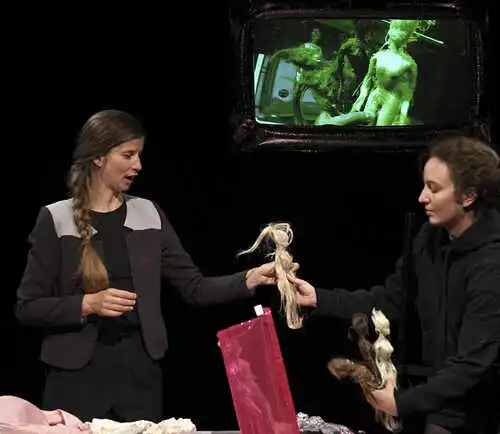 Zwei weiblich gelesene Personen übergeben sich eine Figur aus Haaren. Im Hintergrund ist eine kleine Projektion mit weiteren Haar-Figuren, welche sich auch auf der Bühne befinden.